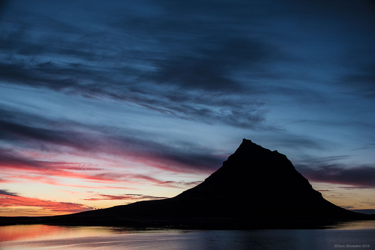 Kirkjufell Mountain stands in silhouette as sunset paints brushstrokes of color over "Church Mountain" and the sea. One of Iceland...